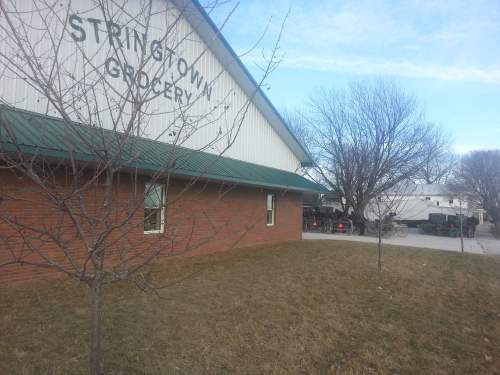 Exterior of Stringtown Grocery with a grassy area and bare trees in the foreground, and vehicles parked nearby.