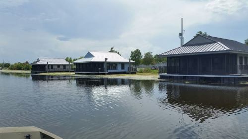Three modern houses on stilts by a calm waterway, surrounded by greenery under a partly cloudy sky.