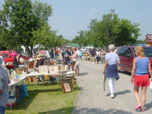 A bustling outdoor flea market with people browsing tables of antiques and collectibles under a clear blue sky.