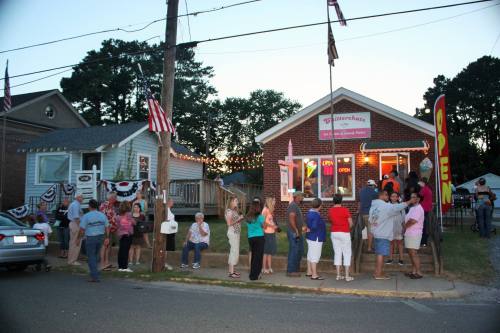 A lively scene outside a small restaurant with a crowd of people waiting in line, decorated with flags and lights.