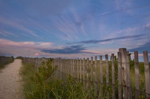 A sandy path leads to a beach, bordered by a wooden fence and lush grass under a colorful sky at dusk.