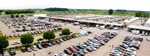 Aerial view of a large parking lot filled with cars, surrounded by buildings and green fields in the background.