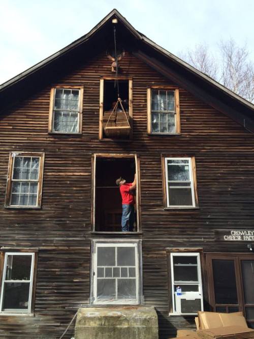 A person stands on a ledge, using a pulley system to lift an object from a window of a rustic, wooden building.