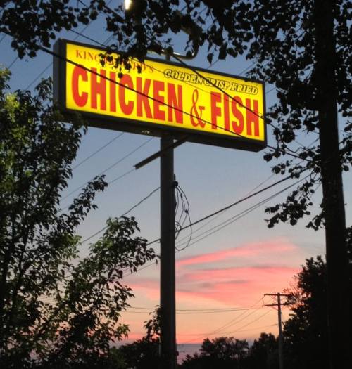 A brightly lit sign for "King's Golden Crisp Inspired Chicken & Fish" against a colorful sunset sky.