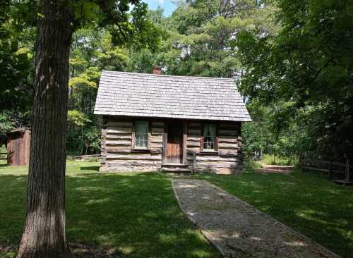 A rustic log cabin with a shingled roof, surrounded by trees and a grassy area, with a stone path leading to the door.