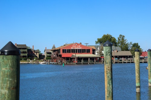 A waterfront restaurant with a red facade, surrounded by trees and buildings, viewed from a calm river with wooden posts in the foreground.