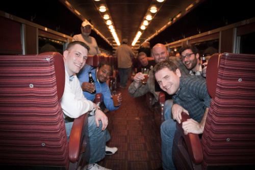 A group of men smiling and posing for a photo inside a train car, with drinks in hand and a festive atmosphere.