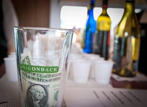 A glass with a dollar bill inside, surrounded by colorful wine bottles and plastic cups on a table.