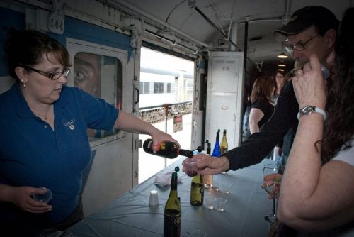 A woman pours wine for a man inside a train car, with bottles and glasses on a table nearby.