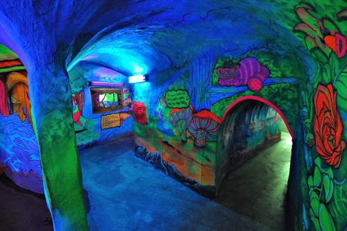 Colorful, graffiti-style murals adorn the walls of a dimly lit, curved tunnel with vibrant blue lighting.