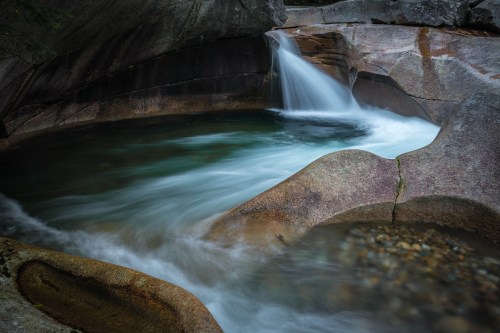 A serene waterfall cascades into a smooth, rocky pool surrounded by textured stone and flowing water.
