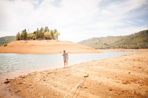 A person stands on a dry, sandy shore near a calm lake, with a small island and trees in the background.