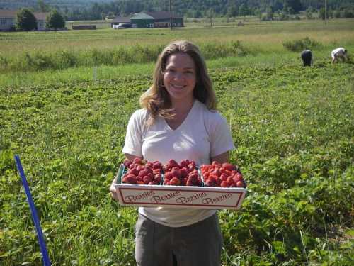 A woman stands in a strawberry field, holding a basket filled with fresh strawberries, with workers in the background.