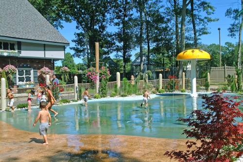 Children playing in a splash pad surrounded by trees and flowers on a sunny day.