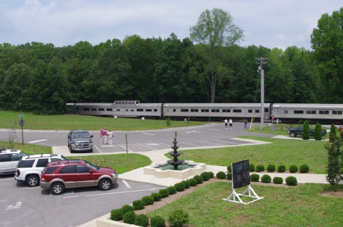 A train passes by a parking area with cars, a fountain, and people walking in a green, wooded setting.