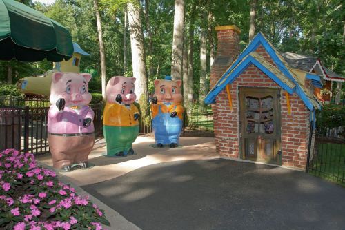 Three colorful pig statues stand outside a small brick house in a park, surrounded by trees and flowers.