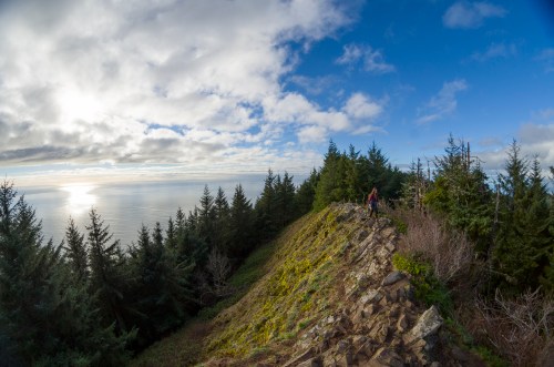 A scenic view of a rocky ridge surrounded by trees, overlooking the ocean under a partly cloudy sky.