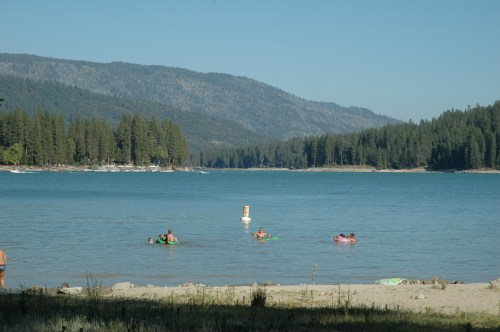 A serene lake scene with people swimming, surrounded by mountains and dense forest under a clear blue sky.