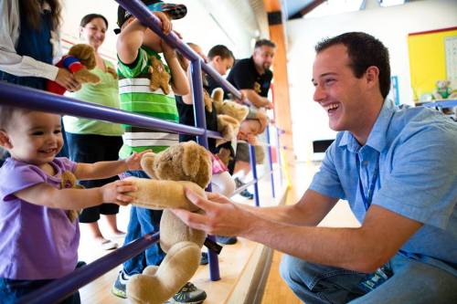 A smiling man hands a teddy bear to a young girl, surrounded by children and adults in a cheerful setting.