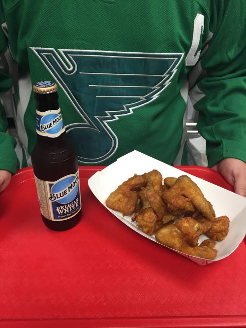 A person in a green jersey holds a tray with a bottle of Blue Moon beer and a serving of fried chicken wings.