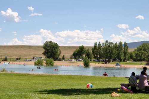 A sunny park scene with a lake, beachgoers, and people relaxing on the grass under a blue sky with fluffy clouds.