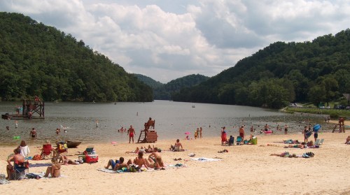 A sandy beach by a lake surrounded by green hills, with people swimming, sunbathing, and enjoying the outdoors.