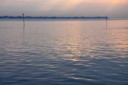 Calm water reflects soft colors of dawn, with distant land and two markers visible in the foreground.