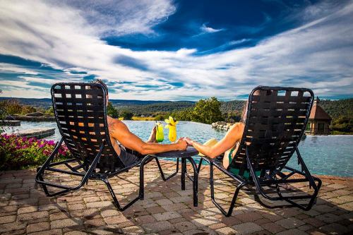 A couple relaxes by a pool, holding hands while sitting in lounge chairs, with a scenic landscape in the background.