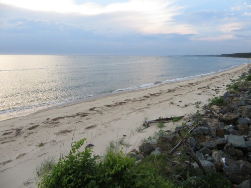A serene beach scene with gentle waves, sandy shore, and distant clouds reflecting on the water under a cloudy sky.