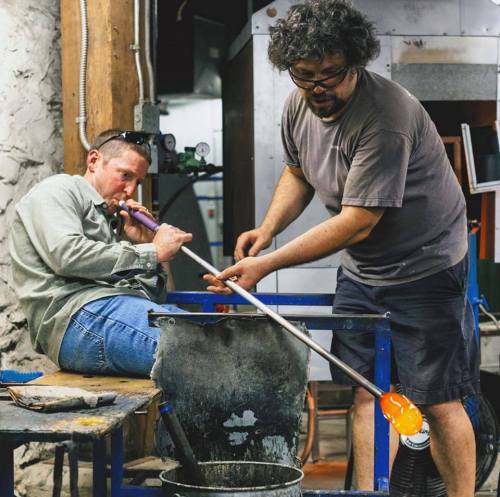 Two glassblowers work together in a studio, one shaping glass while the other sits and plays a flute.