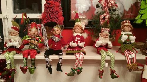 A row of festive elf figurines in colorful outfits, sitting on a shelf decorated for the holidays.