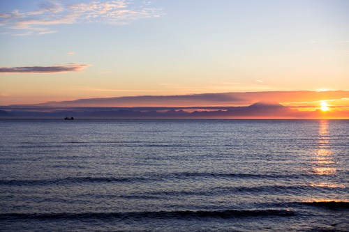 A serene sunset over calm waters, with soft waves and a distant silhouette of a boat on the horizon.