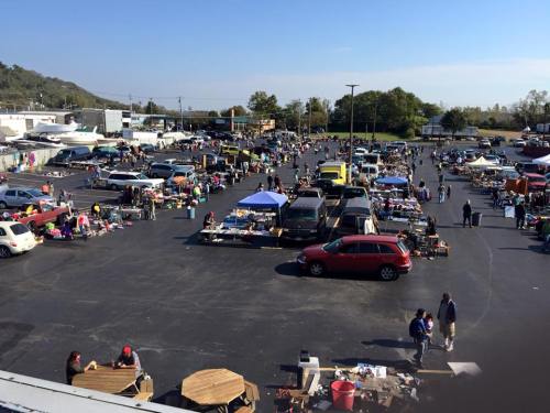 A bustling outdoor flea market with numerous vendors, tents, and shoppers in a large parking lot on a sunny day.