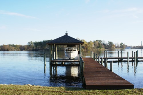 A wooden dock extends into calm water, with a gazebo and a boat moored nearby, surrounded by trees and a clear sky.