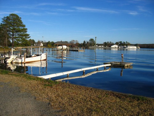A calm lake scene with a wooden dock, boats, and trees under a clear blue sky. Reflections shimmer on the water's surface.