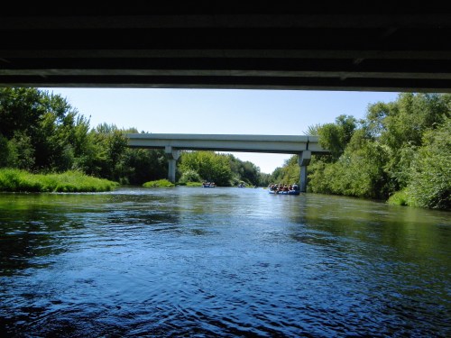A serene river scene viewed from under a bridge, with lush greenery and a few boats in the distance.