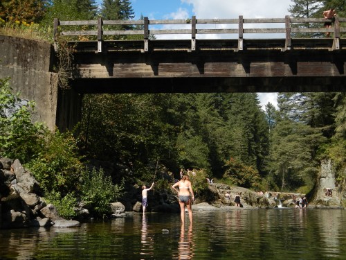 A serene river scene with people fishing and wading under a wooden bridge, surrounded by lush greenery and rocky banks.
