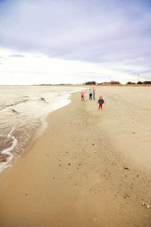 Three children walking along a sandy beach, with gentle waves lapping at the shore under a cloudy sky.