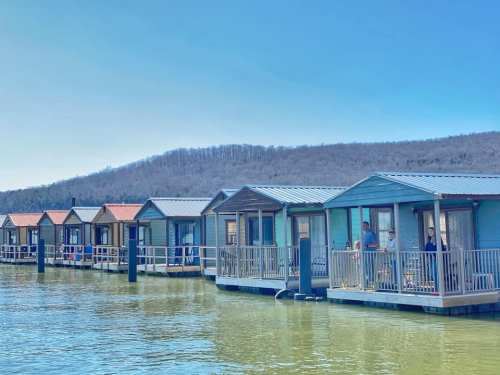 Colorful houseboats lined up on a calm lake, with a backdrop of a wooded hill under a clear blue sky.