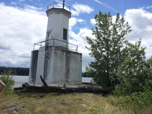 A weathered lighthouse stands by a river, surrounded by greenery and a fallen log under a cloudy sky.