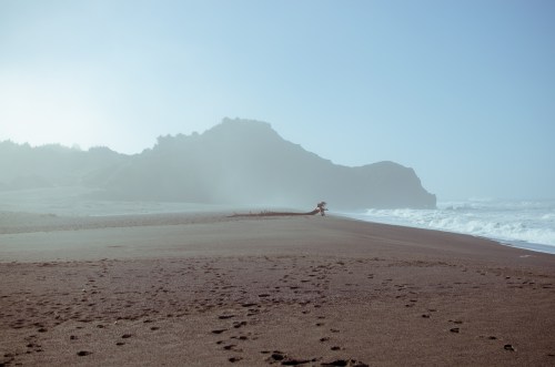 A misty beach scene with gentle waves, footprints in the sand, and a rocky cliff in the background.