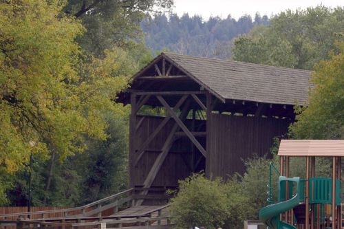 A wooden covered bridge surrounded by trees, with a playground visible in the foreground.