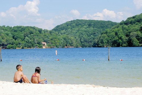 Two children sit on a sandy beach by a calm lake, surrounded by green hills and a clear blue sky.