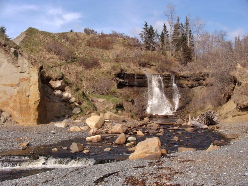 A serene landscape featuring a waterfall cascading down rocky cliffs, surrounded by greenery and a pebbly riverbed.