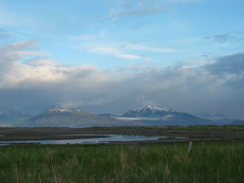 A scenic view of snow-capped mountains under a blue sky, with a calm body of water and green grass in the foreground.