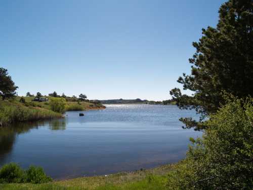 A serene lake surrounded by greenery under a clear blue sky, reflecting sunlight on the water's surface.