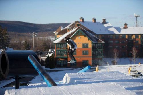 A snowboarder performs a jump over a rail in a snowy terrain park, with a colorful lodge in the background.