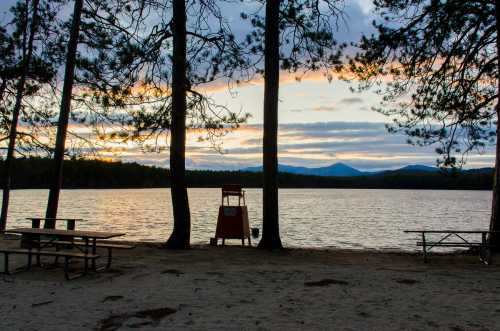 A serene lakeside scene at sunset, framed by trees, with picnic tables and a lifeguard chair by the water.