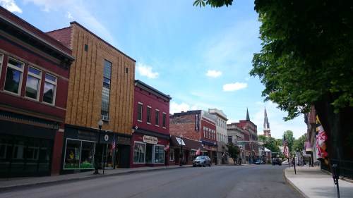 A quiet street in a small town, lined with colorful buildings and a church steeple in the background under a blue sky.