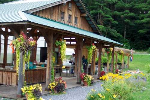 A rustic wooden pavilion surrounded by colorful flowers and hanging plants, with people engaged in activities.